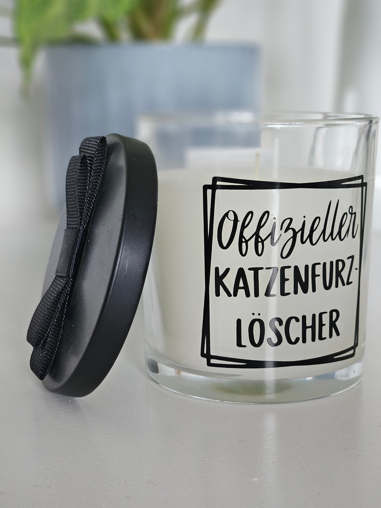 Eine Duftkerze von links Fotografiert, mit der Aufschrift, Offizieller Katzenfurzlöscher. Das Glas ist offen und der Deckel ist angelehnt.