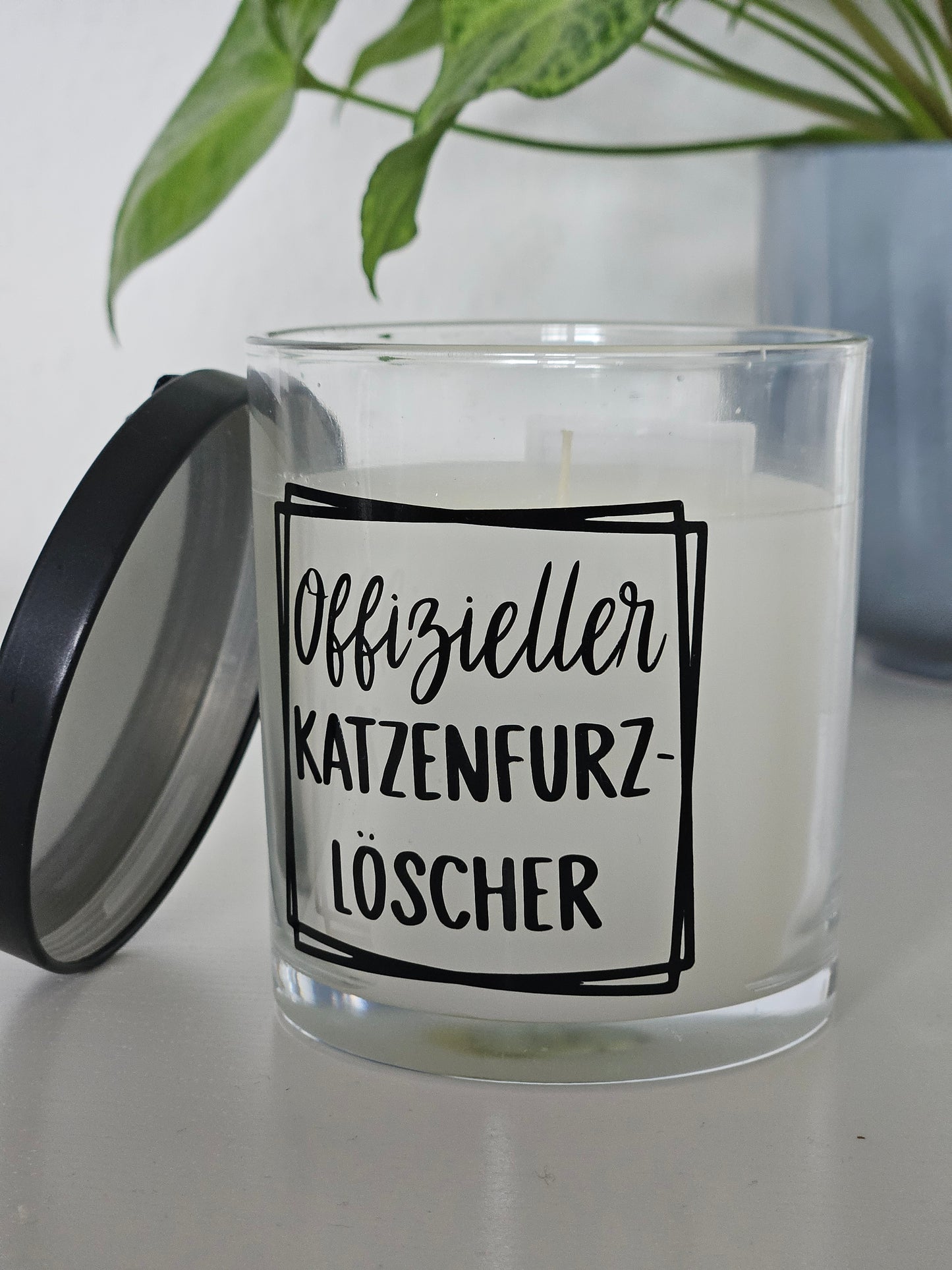 Eine Duftkerze von der Seite fotografiert, mit der Aufschrift, Offizieller Katzenfurzlöscher. Das Glas ist offen und der Deckel ist angelehnt
