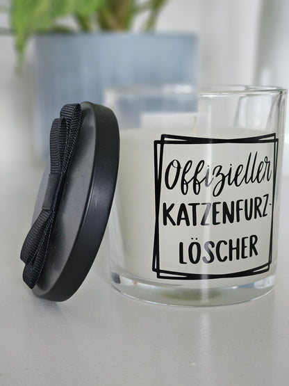 Eine Duftkerze von links Fotografiert, mit der Aufschrift, Offizieller Katzenfurzlöscher. Das Glas ist offen und der Deckel ist angelehnt.